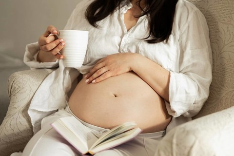 A pregnant woman enjoys a moment of relaxation, sipping tea and reading a book. Her hand gently
