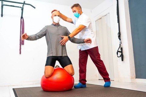 Client balancing on a red ball assisted by Physiotherapist with face mask. Physiotherapy with