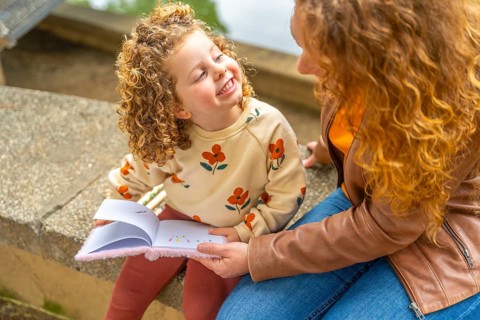 Adorable blonde girl smile while reading a book with her mother outdoors sitting in a park