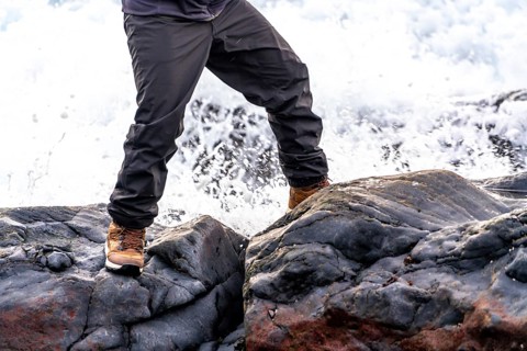A person is standing on a rock near a body of water. The person is wearing a brown jacket and brown