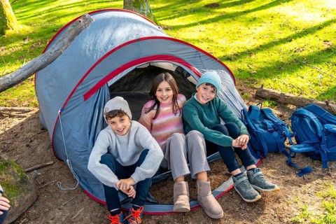 Three caucasian children sitting on a camping tent smiling at camera enjoying a day in the forest