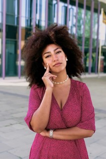A young Arab woman with natural afro hair stands thoughtfully, looking at camera, wearing a pink