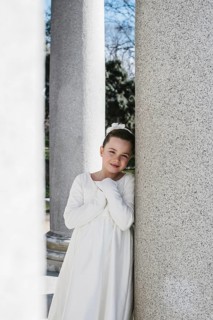 A young girl in a white First Communion dress stands between stone columns, smiling softly. The