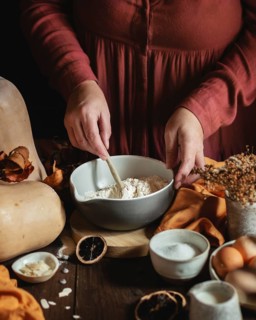 A woman in a cozy kitchen setting mixes flour in a bowl, surrounded by ingredients like eggs,