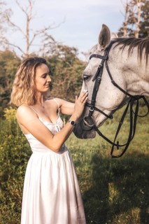 A young woman in a white dress gently touches a horse face, set against a lush green background.
