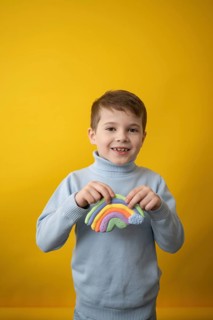 A joyful child in a blue sweater holds a colorful modeling clay rainbow against a bright yellow