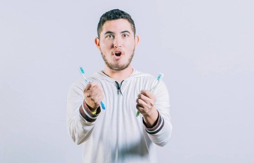 Amazed teen guy holding two toothbrush looking at camera isolated. Surprised people holding