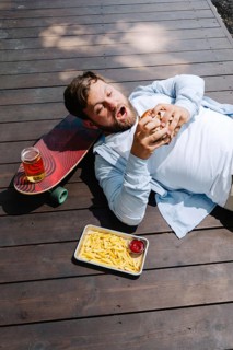 A man enjoys a burger, fries, and a drink while lounging outdoors on a wooden deck. A skateboard