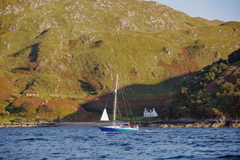 A sailboat sails on water in front of a mountainous landscape with a single house, Mallaig,