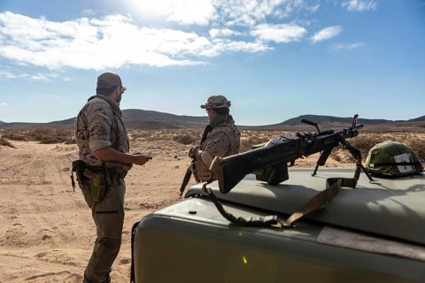 Two soldiers in military gear converse near a vehicle in a desert. A rifle and helmet lie on the