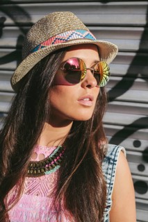 A trendy woman wearing mirrored sunglasses and a woven hat stands against a graffiti wall. Dressed