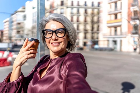 Happy mature woman with gray hair and glasses smiling at camera, holding takeaway coffee cup while