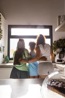 Two women with a child share a warm embrace, facing a sunlit window in a kitchen. This image