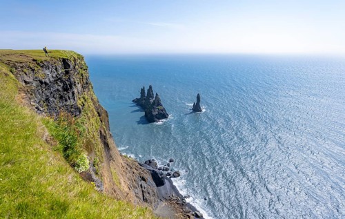 Young man standing on cliff, view from steep cliff to the rocks Reynisdrangar in the water, at