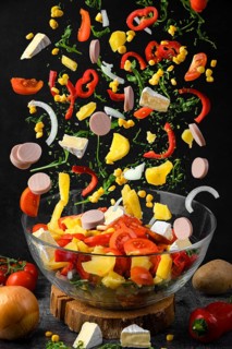 Salad ingredients, such as peppers, tomatoes, sausage, corn, arugula and potatoes fall a glass