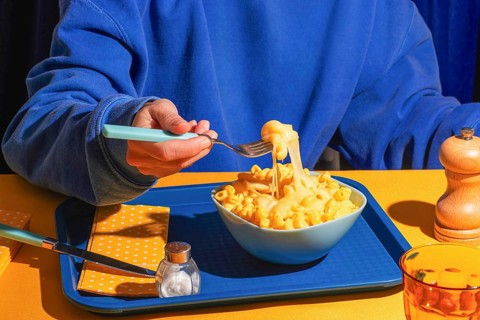 A person in a blue shirt holds a forkful of creamy macaroni and cheese from a bowl. The vibrant