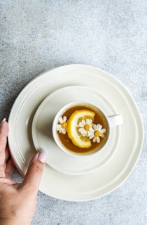 Top view of cropped unrecognizable woman's hands hold a white plate featuring a tea cup with lemon