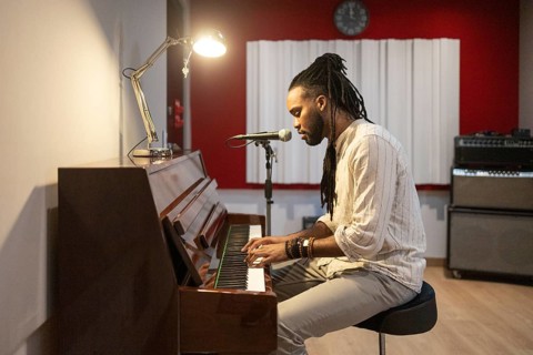 A black musician passionately plays the piano and sings into a microphone in a cozy recording