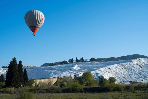 A single hot air balloon flies high above white limestone terraces and trees, sinter terraces, lime