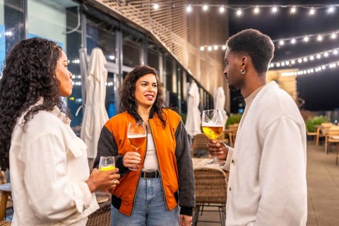 Three multi-ethnic friends drinking beer and talking standing on a bar terrace at night