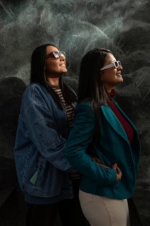 A cheerful Latin lesbian couple wears stylish sunglasses while embracing. Both women smile warmly,