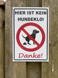 Sign with pictogram and inscription Hier ist kein Hundeklo on the outside of a wooden fence,