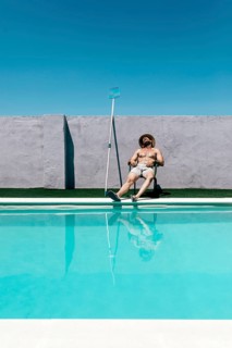 A shirtless man in shorts and a straw hat takes a nap, relaxing on a chair by a clear blue swimming
