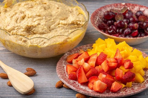 Bowl with dough next to sliced strawberries, mango and grapes for fruitcake