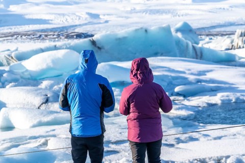 Two people are standing on a frozen lake, looking out at the ice. The man is wearing a blue jacket