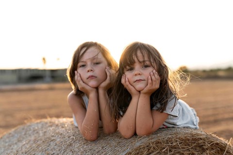 Two young children lie on a hay bale, resting their heads on their hands with a serene expression.