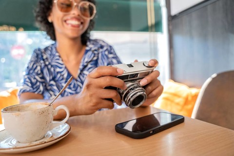 A smiling woman in a patterned blue blouse holds a vintage camera at a cafe. A cup of coffee and a