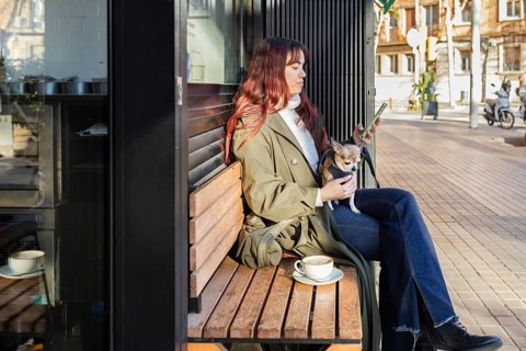 A woman sits on a bench outside a cafe, looking at her phone, with a small chihuahua on her lap.