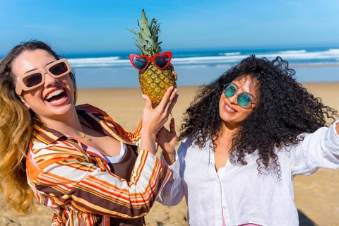 Two young women are smiling and having fun together on a sunny beach, holding a tropical pineapple