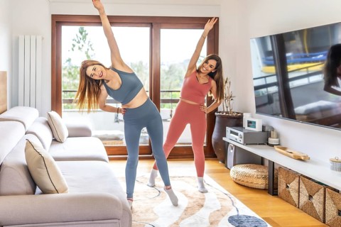 Two smiling women practicing yoga together in their living room, following an online class on their