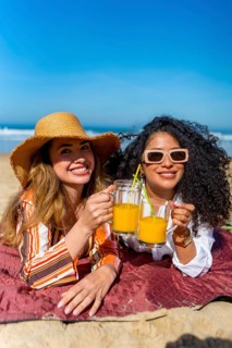 Two smiling friends lying on a blanket at the beach, holding glasses with orange juice, celebrating