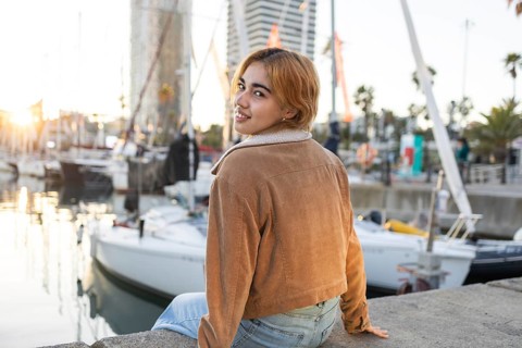 A Mixed-race woman sits by the marina, smiling at the sunset Her casual attire and relaxed demeanor