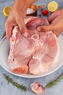 Raw pork shoulder being seasoned with garlic, lemon, salt, and herbs on a white plate. Hands