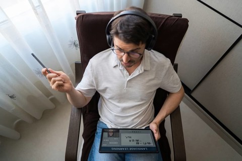A man sits at home with headphones, intently studying Mandarin online using a tablet. The setup