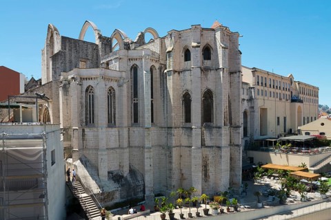The back of a Gothic stone church façade with high windows and ruinous structures in daylight,