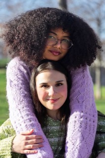 Two friends enjoy a sunny day outdoors, embracing warmly. One wears glasses and a purple sweater,