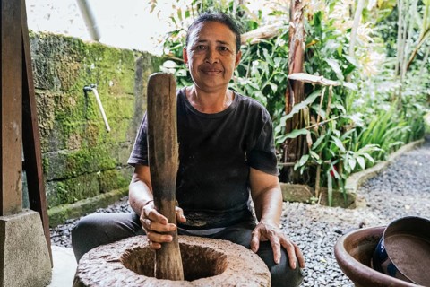 A woman uses a large wooden pestle in a traditional stone mortar for balinese coffee, surrounded by