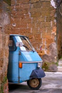 An iconic blue three-wheeled vehicle parked on a quiet, narrow street surrounded by rustic stone