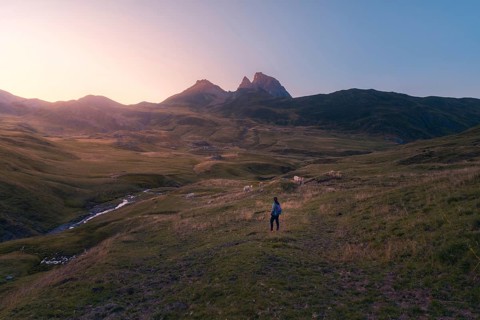 A lone hiker strolls across a vast, serene mountain landscape at sunrise, surrounded by grazing