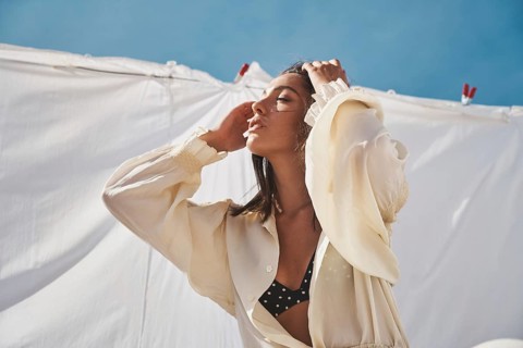 A woman enjoys a sunny day on a rooftop, framed by gently blowing white fabrics She wears a light,