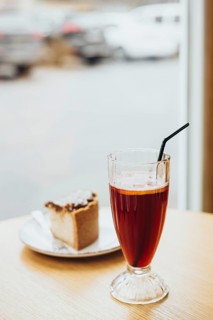A glass of iced tea with a straw and a slice of cake on a wooden table near a window, offering a