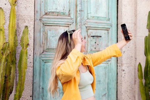 A female tourist wearing a yellow shirt takes a selfie with her smartphone in front of a rustic