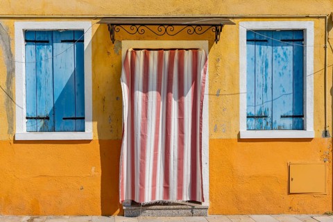 Yellow house facade with entrance door with red and white curtain and windows with blue shutters,