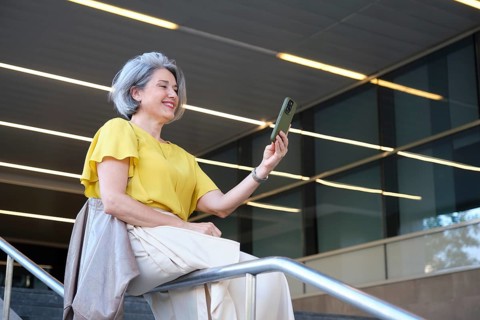 A cheerful mature woman in a yellow blouse takes a selfie with her smartphone while sitting on a