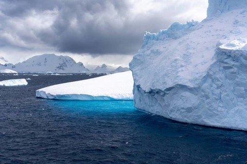 Table icebergs float in the dark Southern Ocean, turquoise underwater edge visible, snow-capped