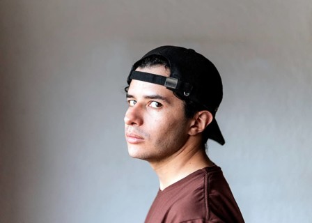 A young male with a neutral expression, wearing a headset and a black cap, poses against a gray
